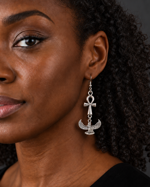 A close-up of a woman wearing a silver dangling earring featuring an Ankh symbol above a winged scarab beetle, set against a dark gray background.