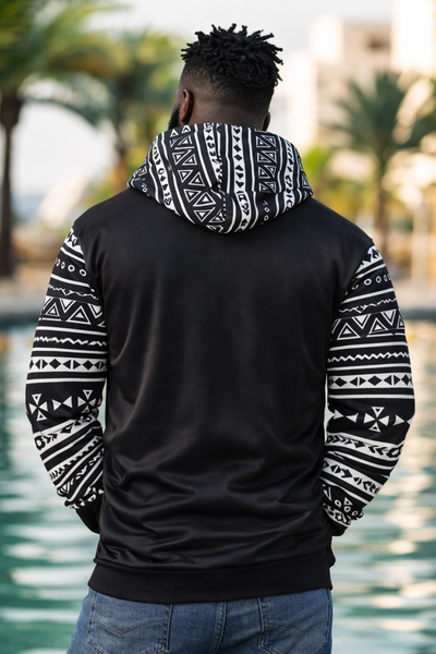 Person wearing a black hoodie with white geometric patterns outdoors by a body of water.