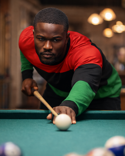Man in a red black and green shirt playing pool in a dimly lit room.