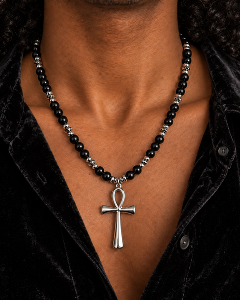 A close-up of a man's neck and chest wearing a beaded necklace with a silver-toned cross pendant over a black velvet shirt.